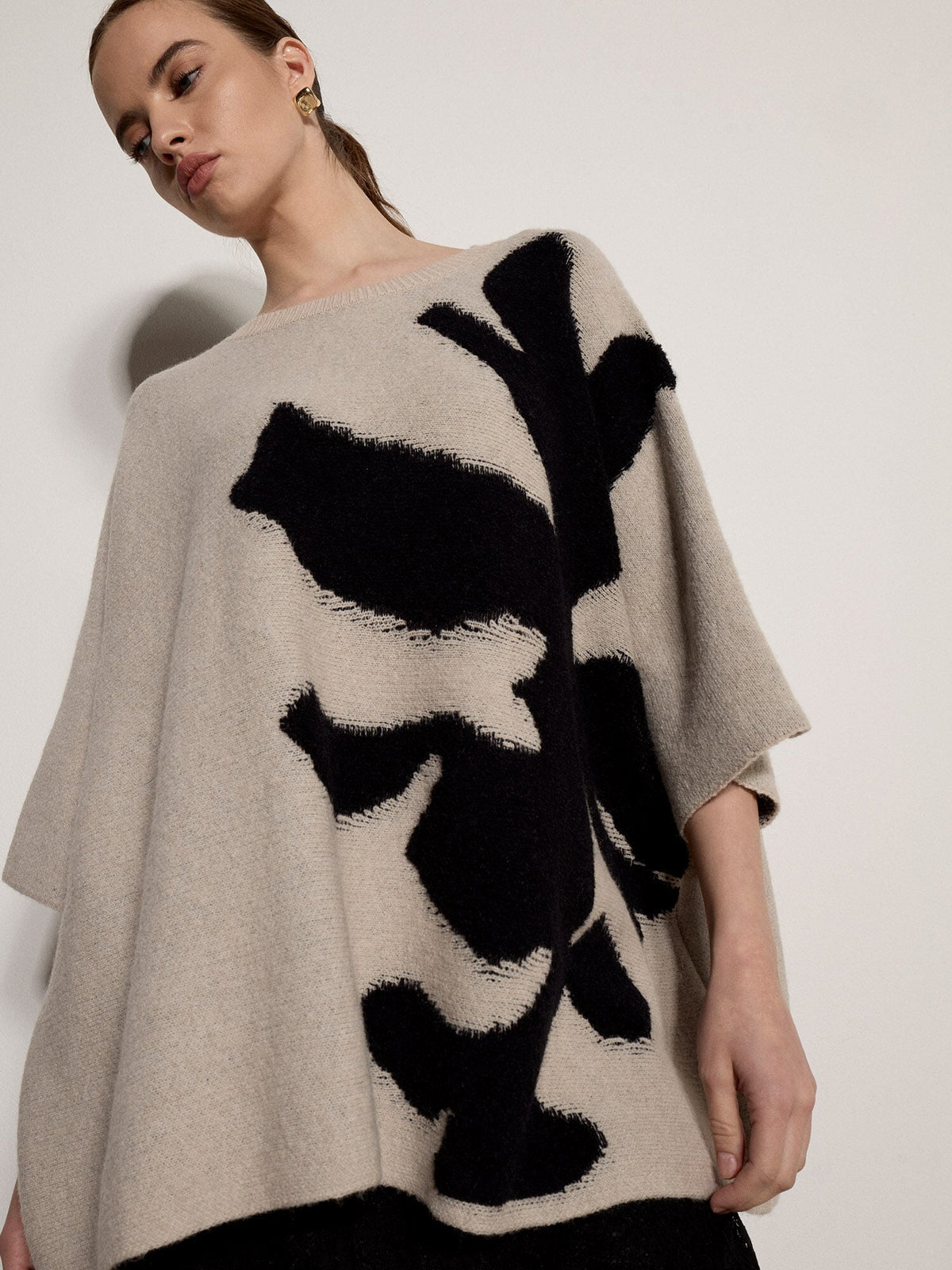 Model wearing oversized knit cape jumper with bold black floral intarsia pattern on stone background