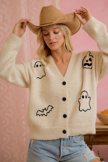 Model wearing ivory button-front cardigan with embroidered Halloween ghost, pumpkin, and bat motifs