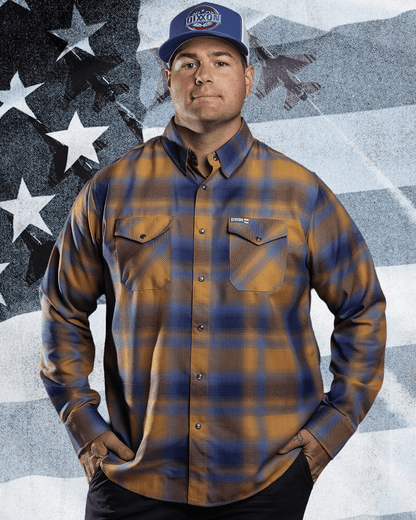 Man wearing Dixxon gold and navy plaid flannel shirt with matching branded cap, hands on hips